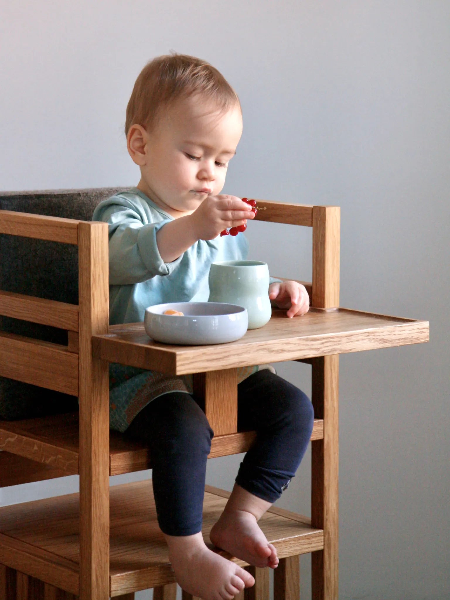 Der ZAUNKÖNIG Lernturm (3-in-1) als Hochstuhl mit Schritthalterung und Tablett aus massivem Eichenholz; Seitenansicht mit Kinder und Kindergeschirr aus Porzellan.