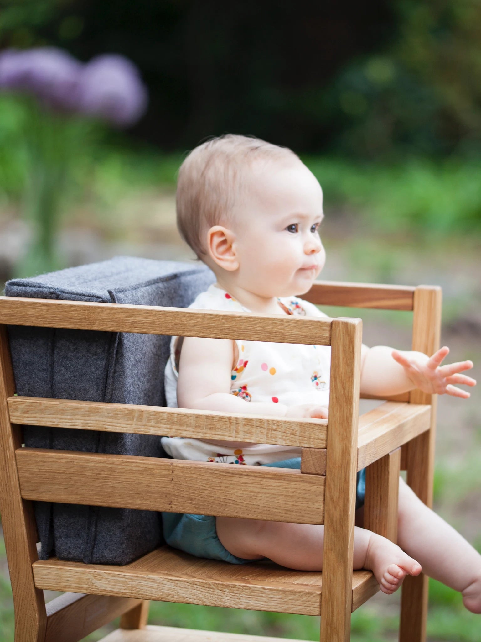 Der ZAUNKÖNIG Lernturm (3-in-1) aus massivem Eichenholz als Hochstuhl im Garten, Seitenansicht mit Kinder, das darin sitzt mit Faltpolster aus Wollfilz "Elephant Grey" als Rückenlehne.