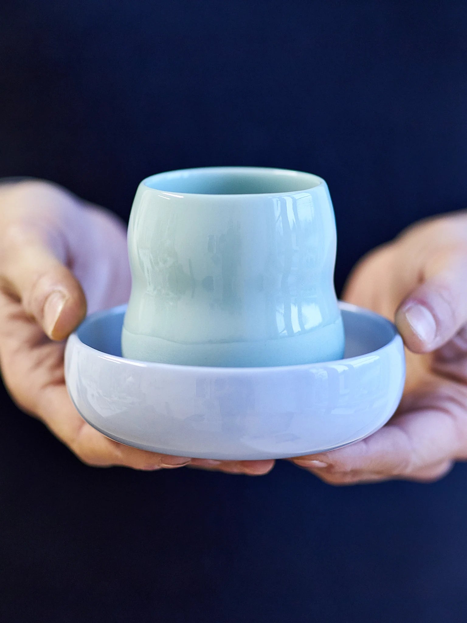 Das Kindergeschirr aus Porzellan von ZAUNKÖNIG, Frontansicht des Kinderbechers in Agave und der Kinderschale in Kornblume in den Händen einer Erwachsenen.