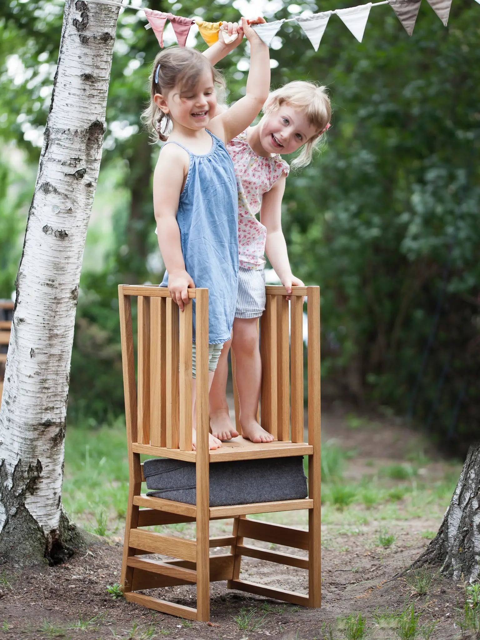 Der ZAUNKÖNIG Lernturm (3-in-1) aus massivem Eichenholz im Garten mit Faltposter aus Wollfilz; Frontansicht mit zwei Kindern, die darauf stehen.