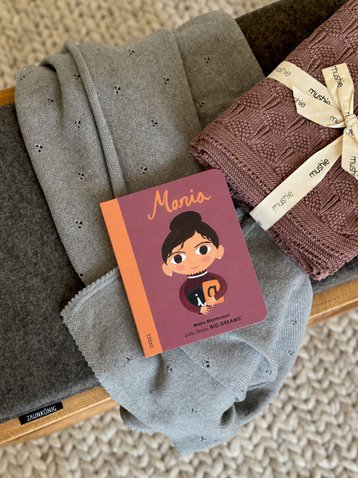 Ein Kinderbuch über Maria Montessori liegt auf dem ZAUNKÖNIG® als Sitzbank mit Kinderdecken von Mushie