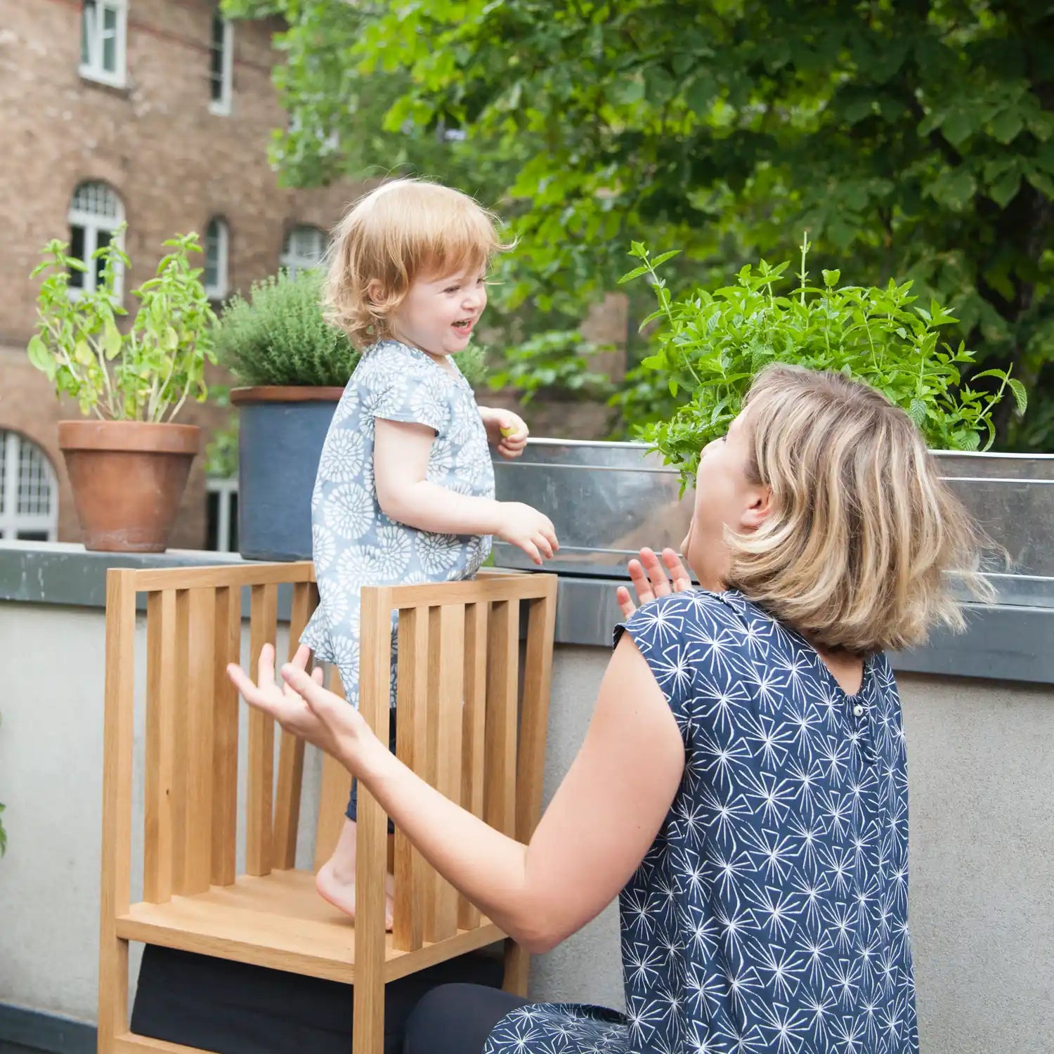 Der ZAUNKÖNIG Lernturm (3-in-1) aus massivem Eichenholz auf dem Balkon, Seitenansicht mit Kind und Mutter. 